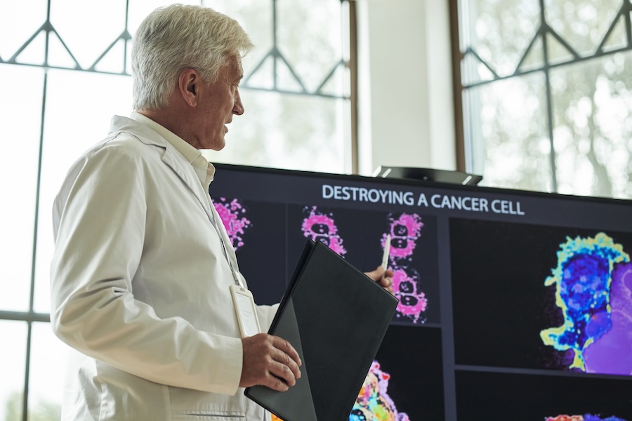 Senior Caucasian man presenting scientific research, holding folder and gesturing toward screen displaying microscopic images and text about destroying cancer cell in laboratory setting